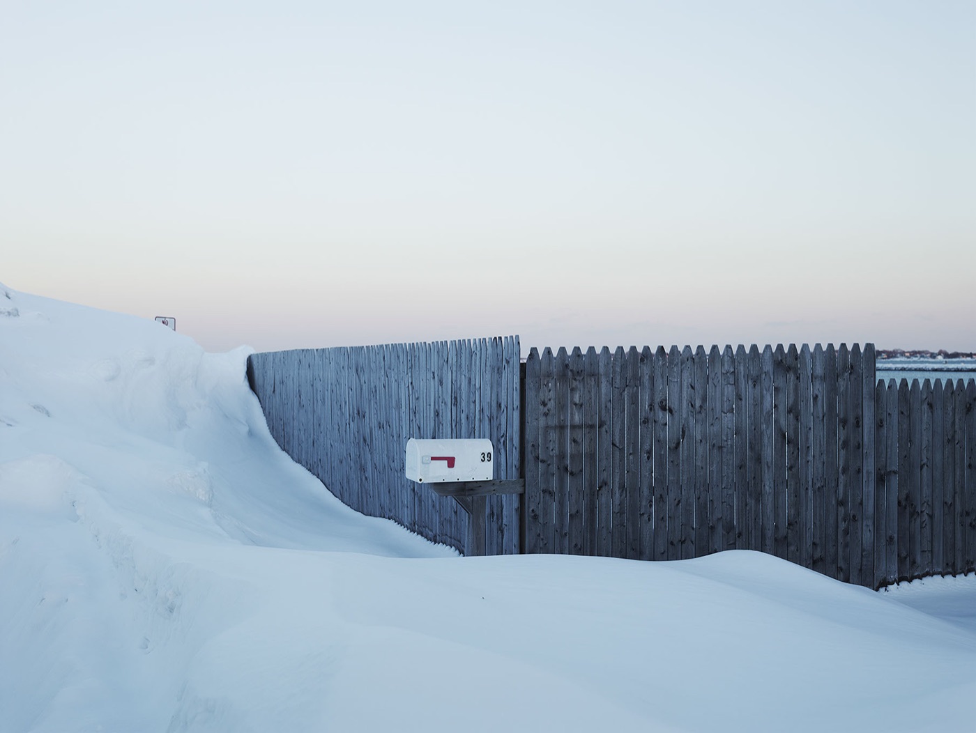 A white mailbox on a weathered fence in deep snow at dusk