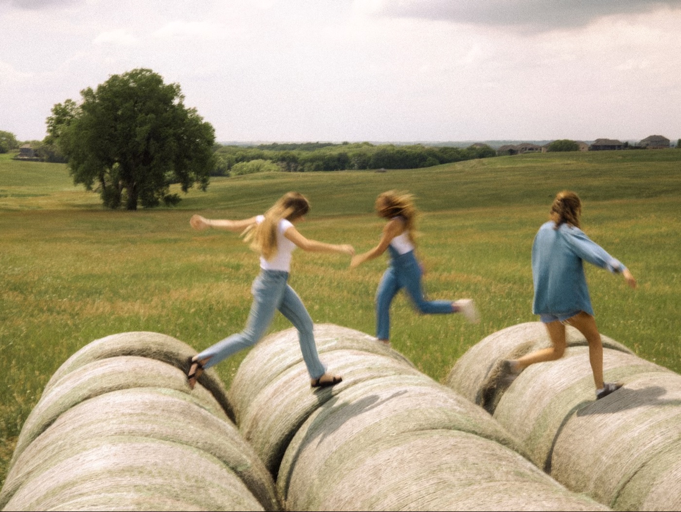 Three girls running and leaping across round hay bales in an open field