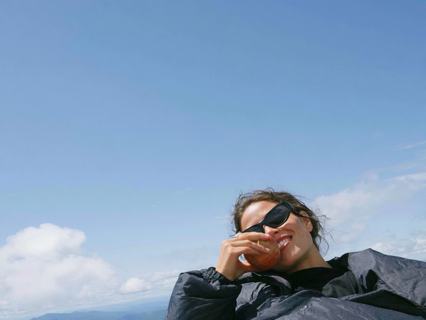 A woman in a rain jacket and sunglasses biting into an apple on a mountain summit